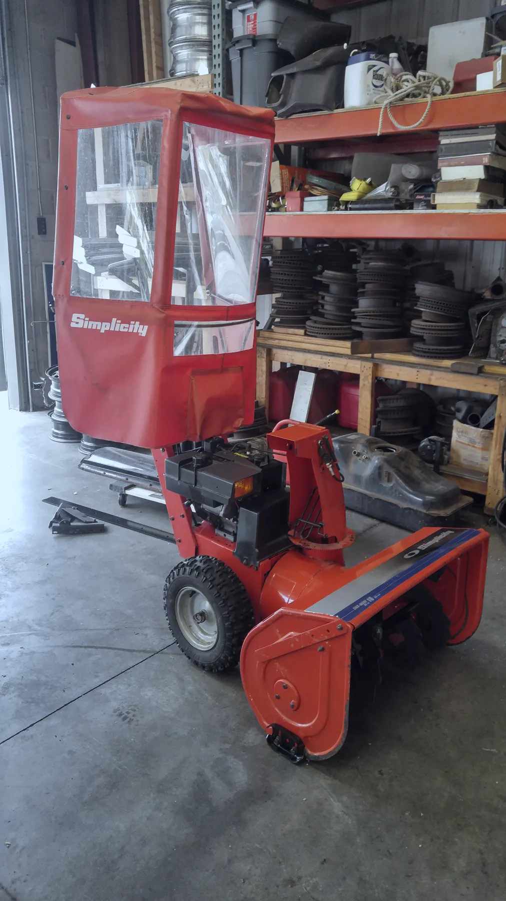 Red snowblower with an enclosed cab beside shelves of brake rotors